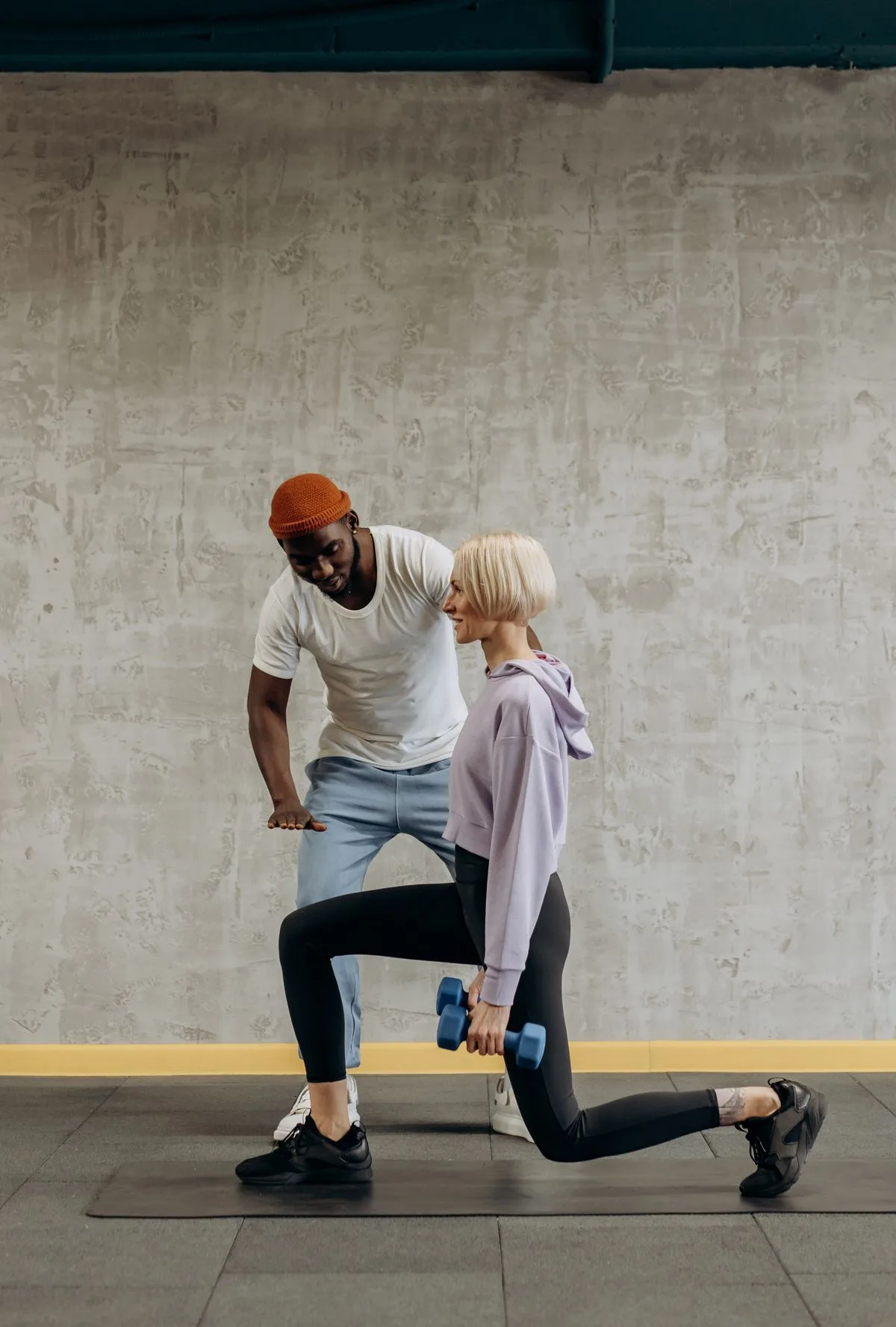Un coach observe une pratiquante qui réalise une fente avec des haltères dans une salle.